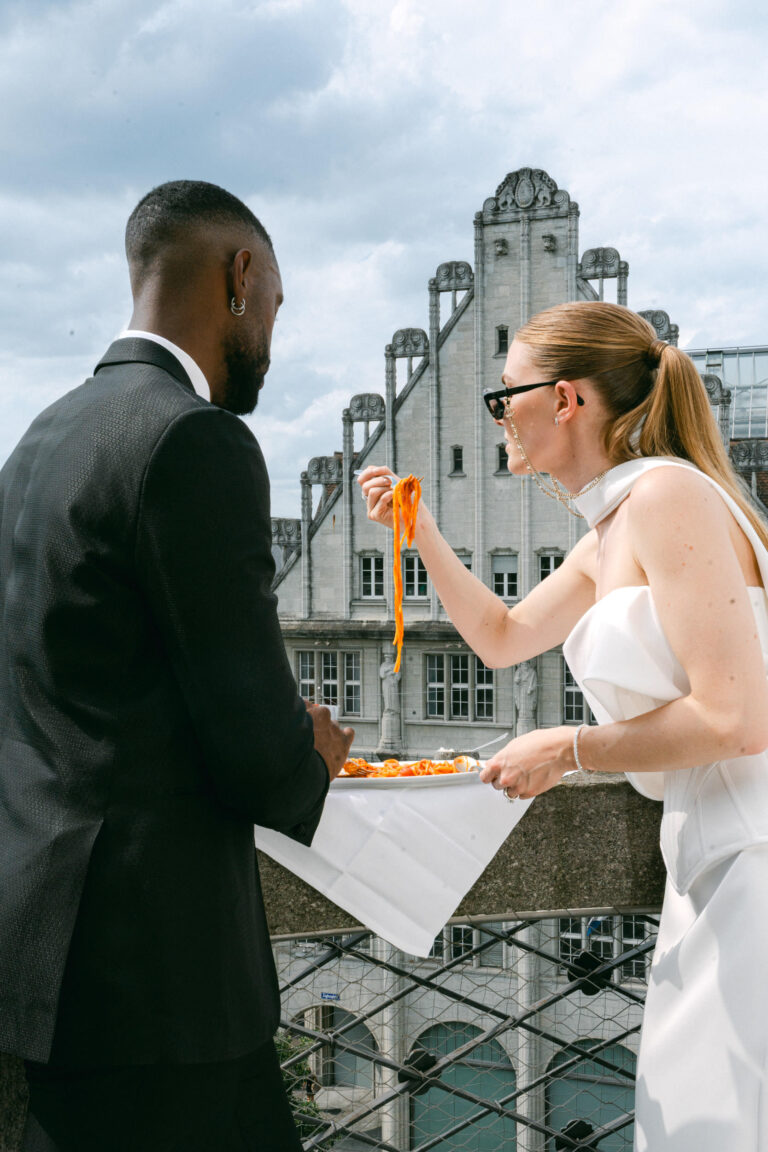 Eine Frau im weißen Kleid und ein Mann im Anzug genießen gemeinsam Pasta auf einem städtischen Dach mit historischen Gebäuden im Hintergrund - ein eleganter Moment, kuratiert von einem Hochzeitsplaner in der Schweiz.