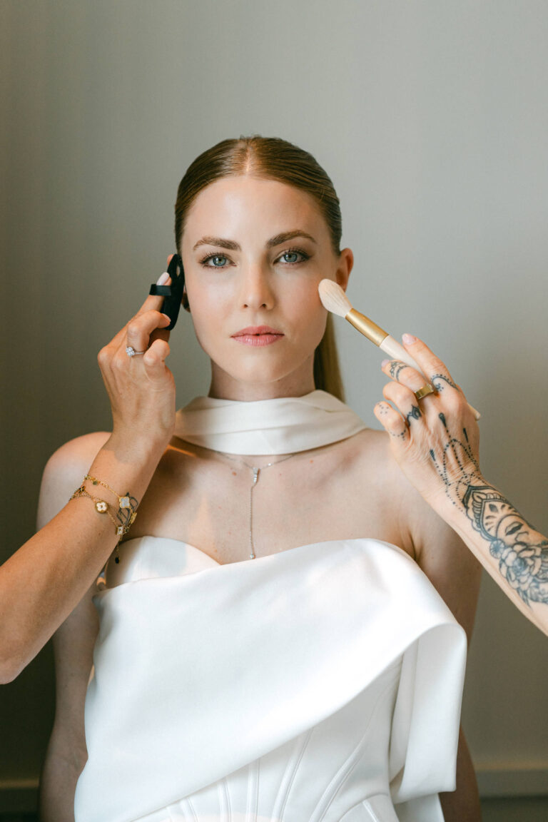 Bei der Hochzeitsplanung in der Schweiz wird einer Frau im weißen Kleid von zwei Stylisten Make-up mit Pinsel und Schwamm aufgetragen.