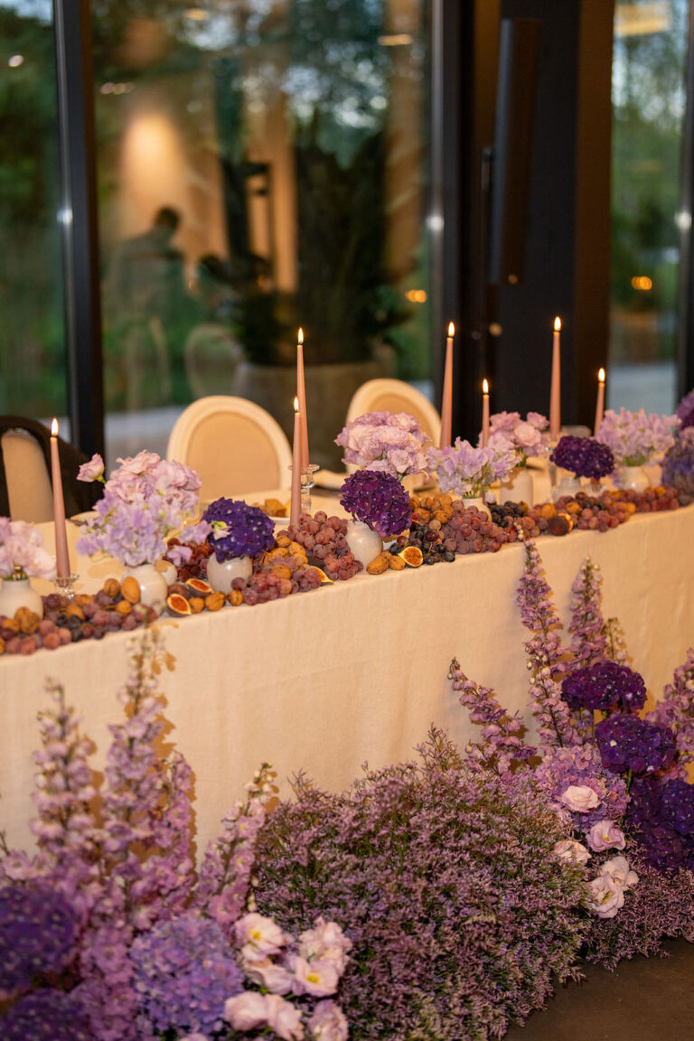 Ein eleganter Tisch mit violetten Blumen, Weintrauben, Kerzen und cremefarbenen Stühlen vor großen Fenstern mit Grünpflanzen im Freien - perfekt für einen Hochzeitsplaner, der Inspiration für verträumte Feiern in der Schweiz oder Italien sucht.