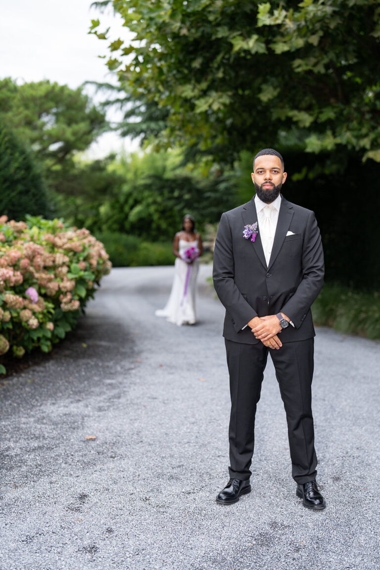 Ein Bräutigam in einem schwarzen Anzug steht auf einem Weg, während seine Braut mit Blumen in der Hand verschwommen im Hintergrund steht - eine elegante Szene, die die Romantik der Hochzeitsplanung in Italien einfängt.