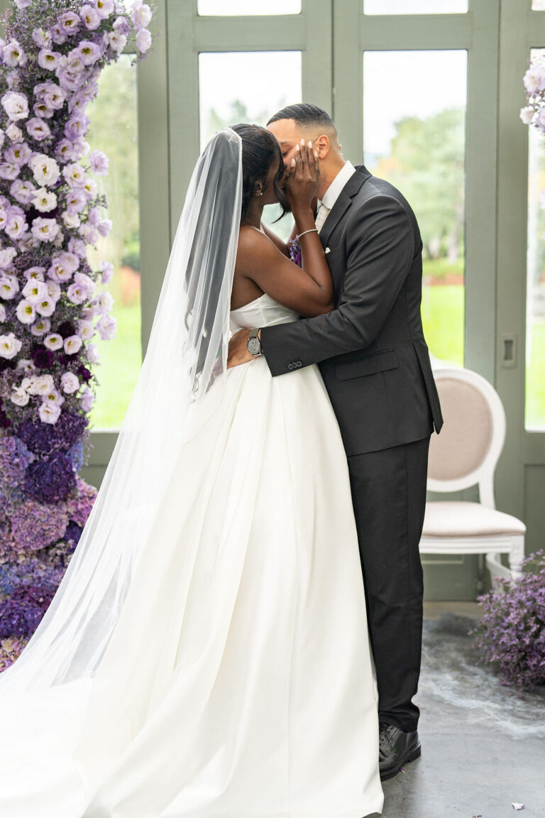 Braut und Bräutigam küssen sich bei ihrer Indoor-Hochzeitszeremonie, umgeben von violetten Blumen - ein eleganter Moment, wunderschön arrangiert von einem Hochzeitsplaner aus der Schweiz oder Italien.