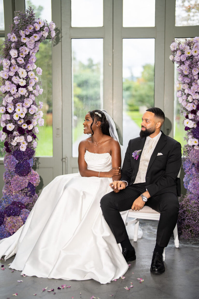 Braut und Bräutigam sitzen lächelnd, händchenhaltend und umgeben von lila Blumenarrangements vor hohen Fenstern - ein bezaubernder Moment, den die erfahrene Hochzeitsplanung für eine Traumhochzeit in Italien ins Leben gerufen hat.