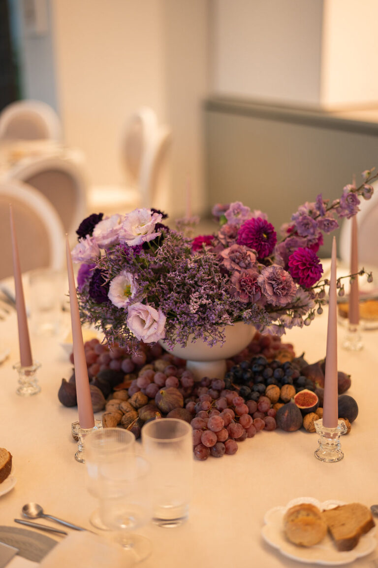 Ein Tafelaufsatz aus violetten Blumen und Trauben, umgeben von Spitzkerzen auf einem gedeckten Esstisch - die perfekte Inspiration für die Hochzeitsplanung in der Schweiz oder Italien.