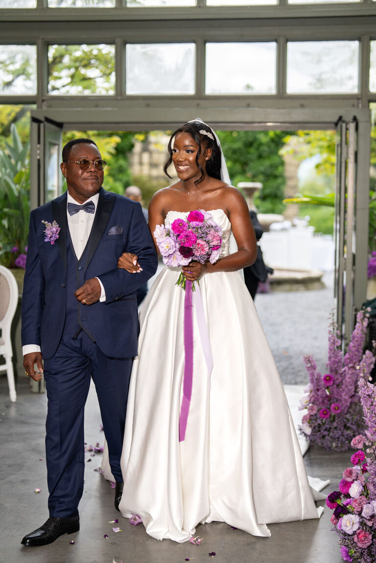 Eine Braut in einem weißen Kleid und mit Blumen in der Hand schreitet mit einem Mann in einem marineblauen Anzug zum Altar und zeigt damit elegante Hochzeitsplanung vom Feinsten.