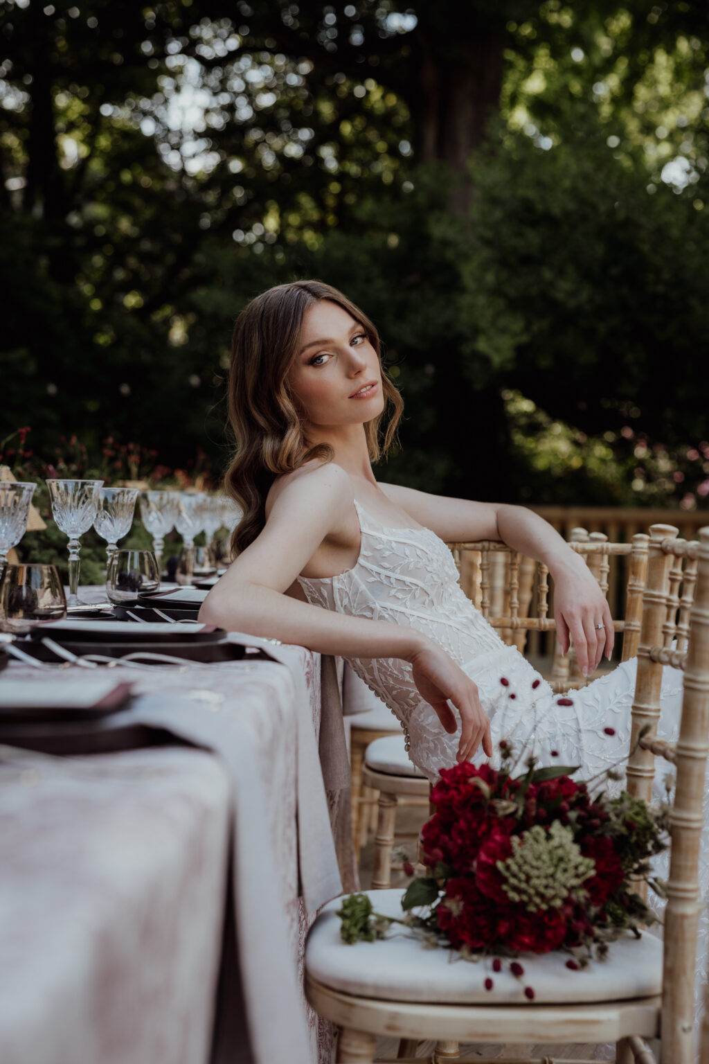 Home 9 Eine Frau in einem weißen Kleid sitzt auf einem Stuhl im Freien an einem gedeckten Tisch, in der Nähe ein Strauß roter Blumen - die perfekte Inspiration für die Hochzeitsplanung in der Schweiz oder Italien.