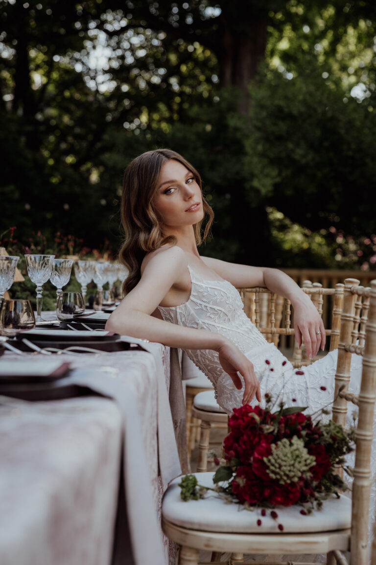 Eine Frau in einem weißen Kleid sitzt auf einem Stuhl im Freien an einem gedeckten Tisch, in der Nähe ein Strauß roter Blumen - die perfekte Inspiration für die Hochzeitsplanung in der Schweiz oder Italien.