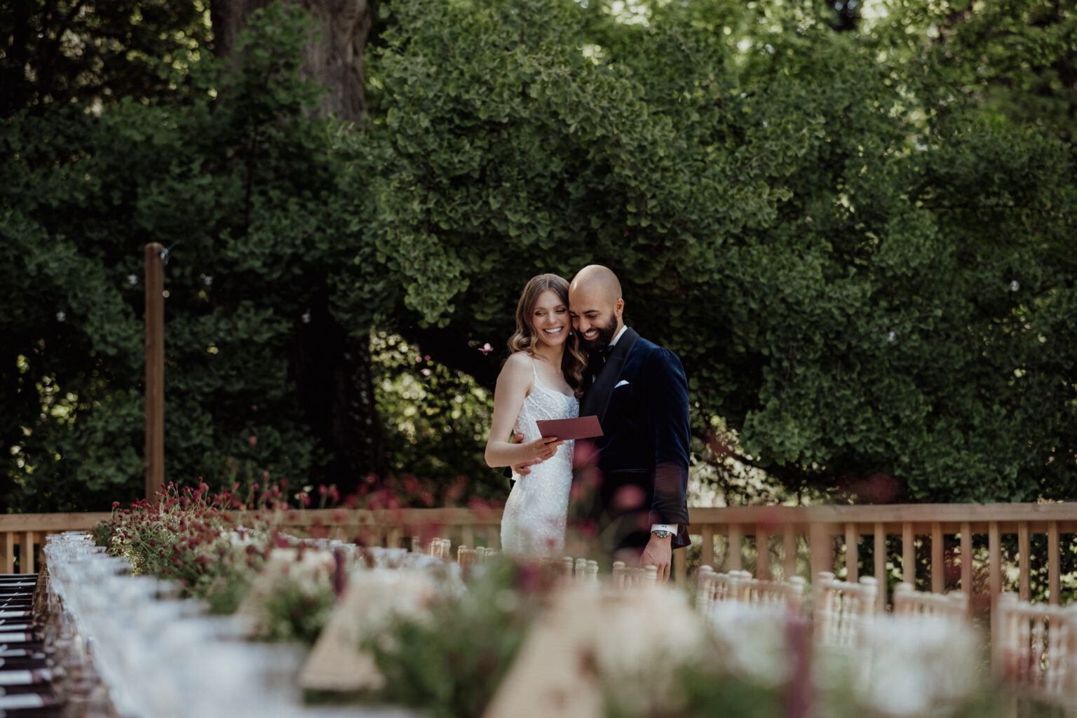 Home 7 Ein Paar umarmt sich und liest gemeinsam eine Karte bei einem Hochzeitsempfang im Freien mit üppigem Grün im Hintergrund, der von einer talentierten Hochzeitsplanerin wunderschön organisiert wurde.