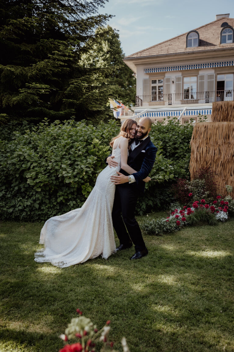 Eine Braut und ein Bräutigam umarmen sich freudig lächelnd in einem Garten, mit einem Haus im Hintergrund - perfekt für alle, die von italienischem Flair oder einer nahtlosen Hochzeitsplanung träumen.