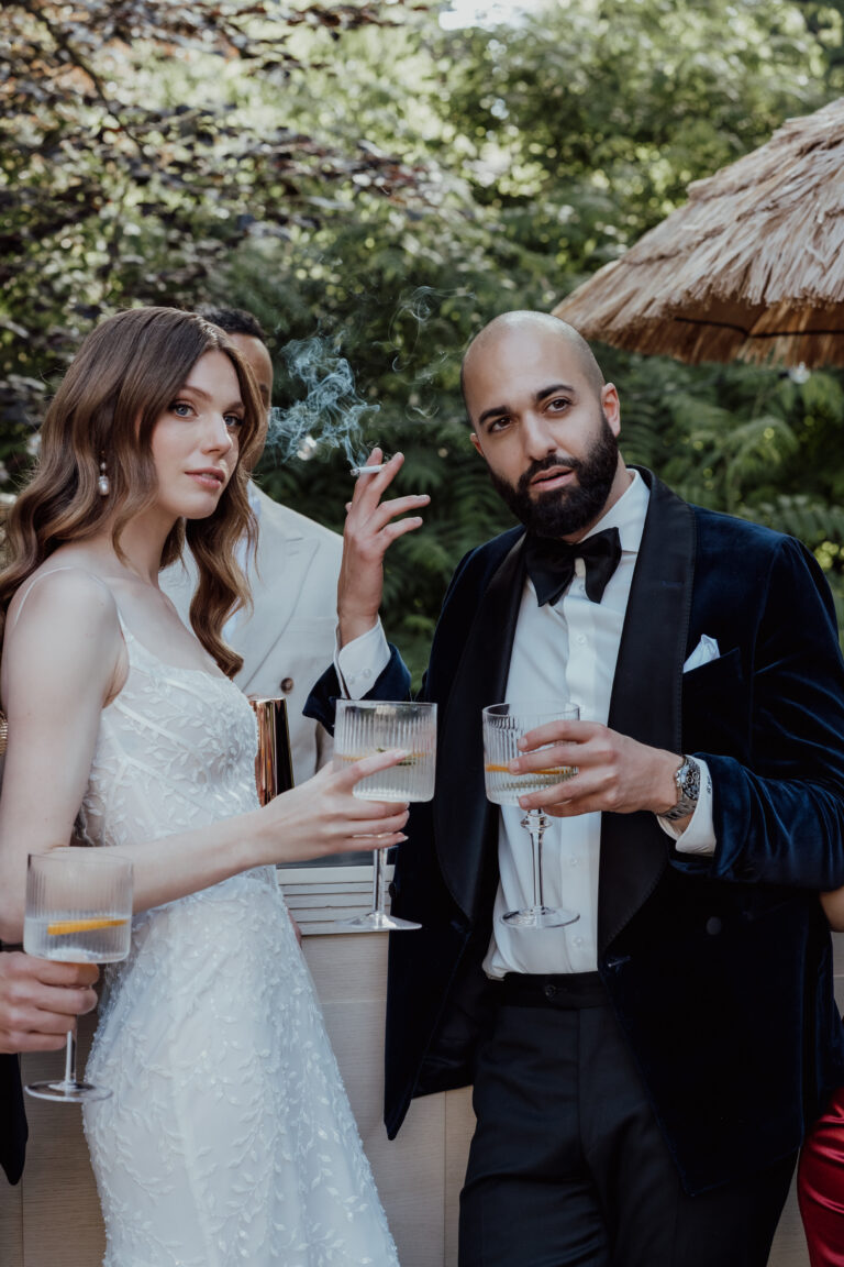 Eine Frau im weißen Kleid und ein Mann im Smoking halten Drinks in der Hand und posieren im Freien bei einer formellen Veranstaltung, die von einer erfahrenen Hochzeitsplanerin für eine elegante, von der Schweiz und Italien inspirierte Feier gestylt wurde.