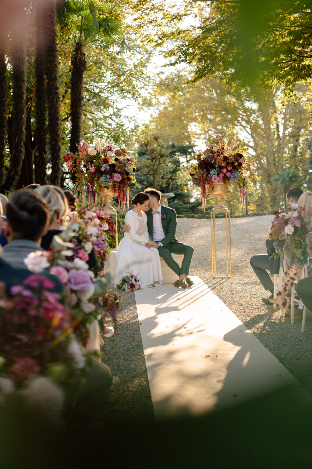 Home 23 Die Braut und der Bräutigam sitzen bei einer Trauung im Freien in Italien zusammen, umgeben von Gästen und farbenfrohen Blumenarrangements, die vom Hochzeitsplaner perfekt aufeinander abgestimmt wurden.