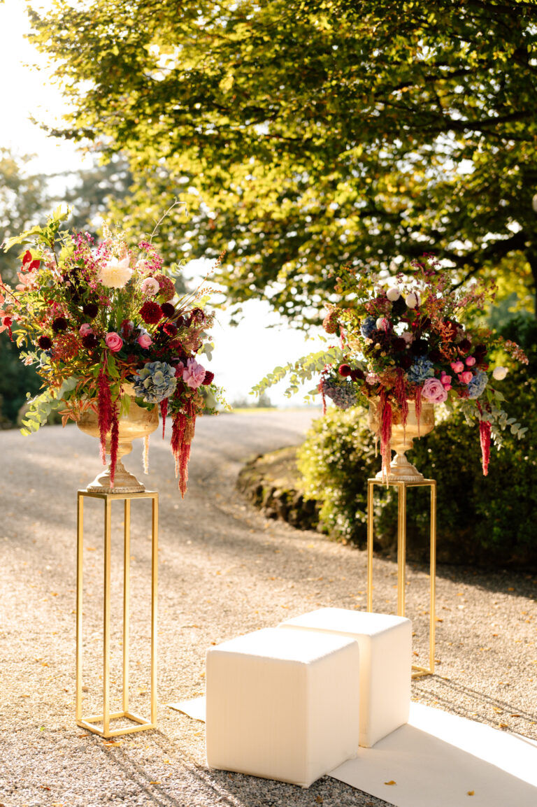 Zwei weiße Bänke und hohe Blumenarrangements sitzen im Freien auf einem sonnenbeschienenen Weg, umgeben von Grün - die perfekte Inspiration für italienische Hochzeitsplaner, die auf der Suche nach verträumten Ideen für die Hochzeitsplanung sind.