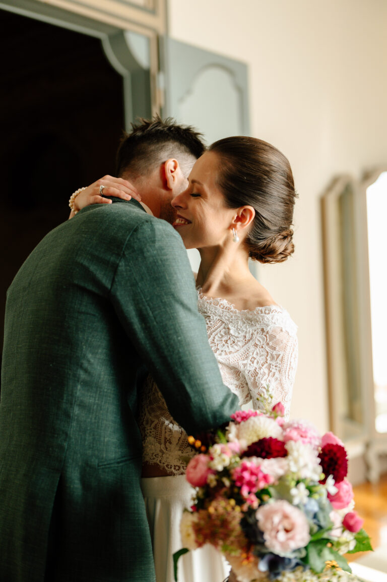 Braut und Bräutigam umarmen sich in einem Raum, die Braut lächelt und hält einen bunten Blumenstrauß in der Hand - ein eleganter Moment, wunderschön arrangiert von einem professionellen Hochzeitsplaner in der Schweiz.