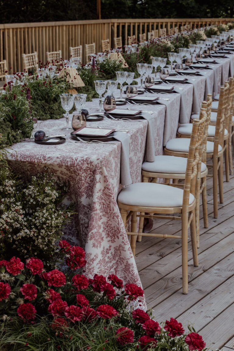 Eleganter Esstisch im Freien mit Blumentischdecken, Gläsern und roten Blumen auf der Holzterrasse - perfekt für eine Hochzeitsplanung, die vom Charme Italiens oder der Schönheit der Schweiz inspiriert ist.