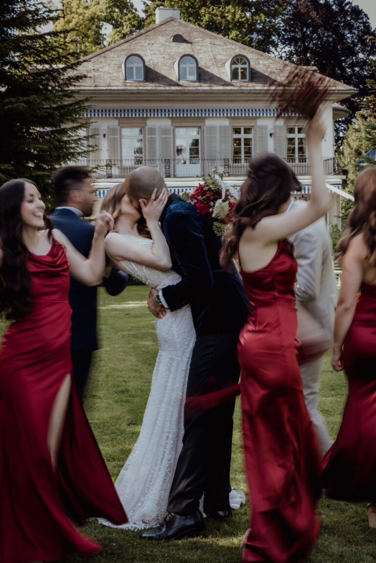 Ein Paar küsst sich vor einem Haus, während die Brautjungfern in roten Kleidern um sie herum feiern und einen magischen Moment der Hochzeitsplanung in der Schweiz einfangen.