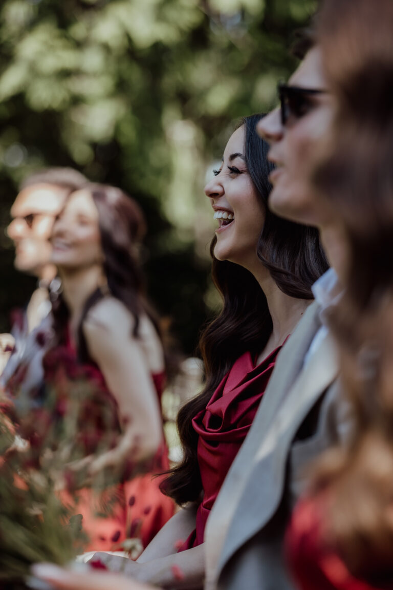 Frauen in roten Kleidern und ein Mann mit Sonnenbrille lächeln im Freien in einer sonnigen, grünen Umgebung - die perfekte Inspiration für die Hochzeitsplanung in der Schweiz.