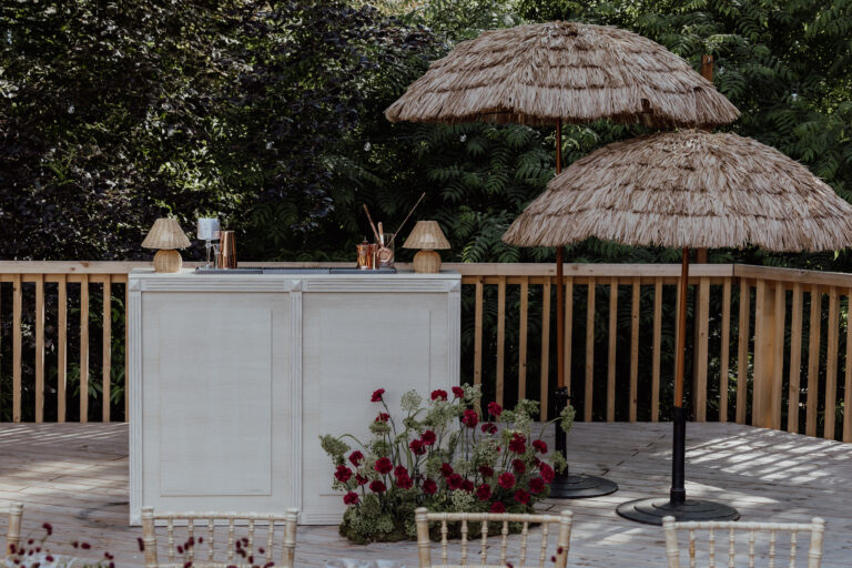 Eine Bar im Freien mit Strohlampen und Sonnenschirmen, roten Blumen am Sockel, umgeben von Grünpflanzen und einem Holzgeländer - perfekt für eine italienische Hochzeitsplanung oder die Vision eines verträumten Hochzeitsplaners.