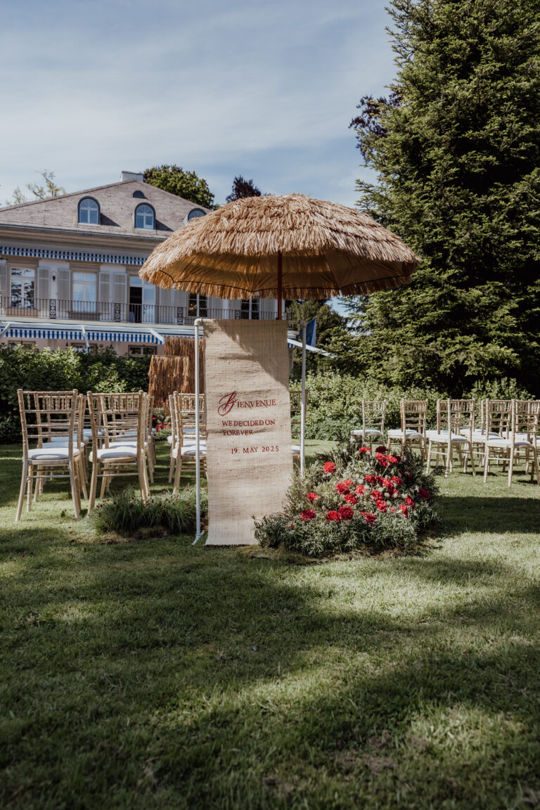 Hochzeit im Freien in Italien mit Holzstühlen, einem strohgedeckten Schirm und einem Willkommensschild auf einer Wiese - die perfekte Inspiration für Ihre Hochzeitsplanung.