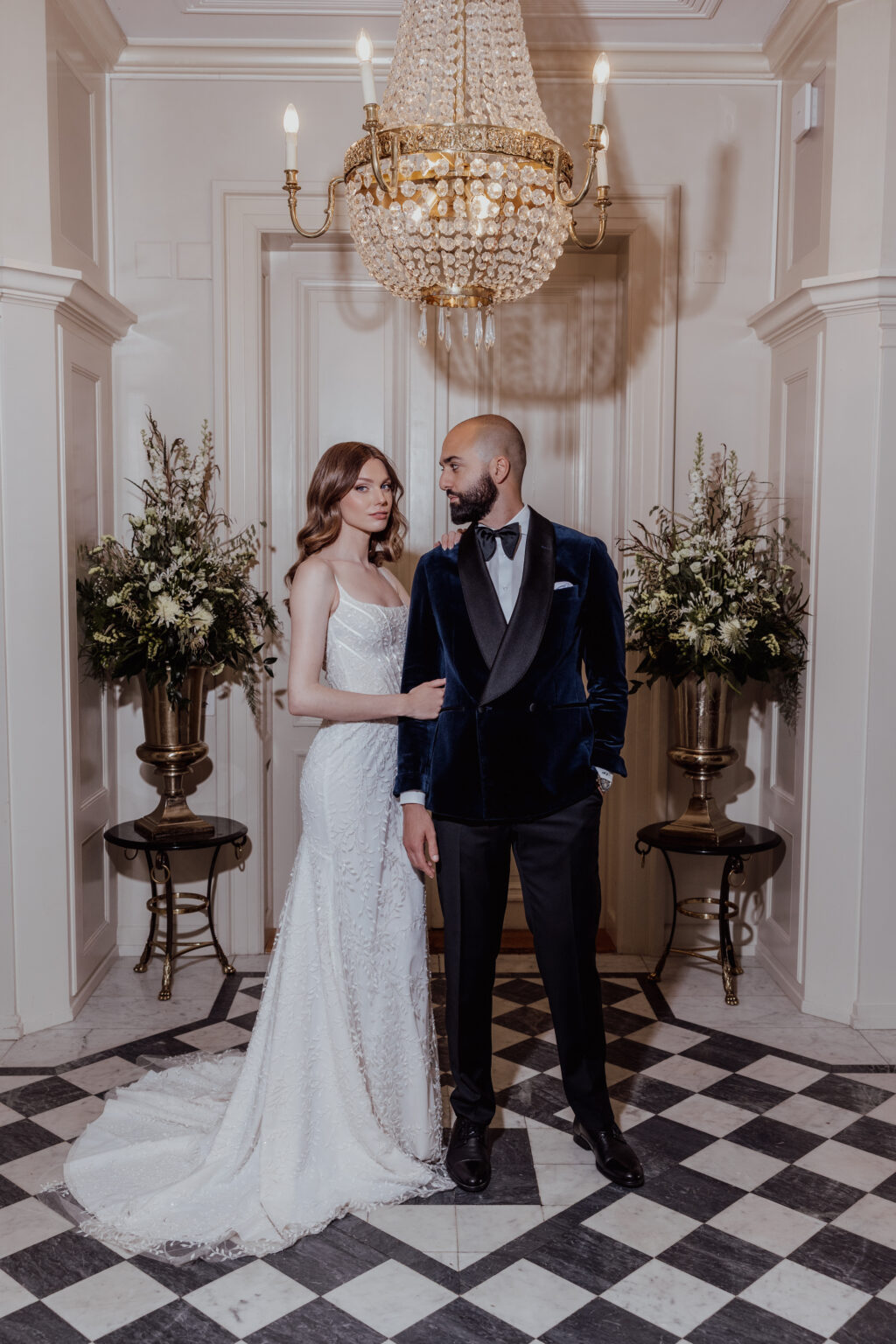Home 15 Die Braut im weißen Kleid und der Bräutigam im Samt-Smoking stehen gemeinsam unter einem Kronleuchter in einem eleganten Flur und zeigen eine exquisite Hochzeitsplanung, die vom raffinierten Stil der Schweiz inspiriert ist.