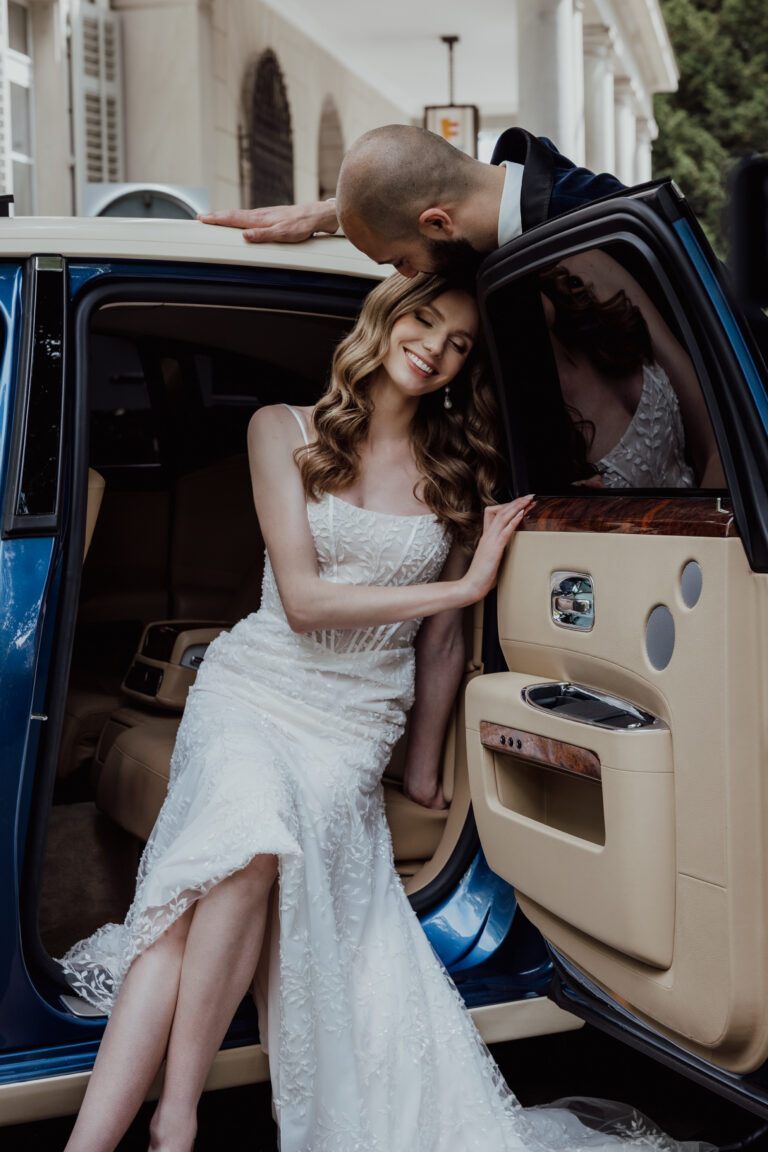Eine Braut in einem weißen Kleid lächelt, als ihr Bräutigam, begleitet von einem professionellen Wedding Planner, sie in einem Luxusauto mit offener Tür in der schönen Schweiz auf den Kopf küsst.