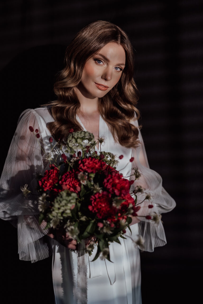Eine Frau in einem weißen Kleid mit einem Strauß roter und weißer Blumen in dramatischem Licht - die perfekte Inspiration für einen italienischen Hochzeitsplaner oder ein Brautpaarshooting in der Schweiz.