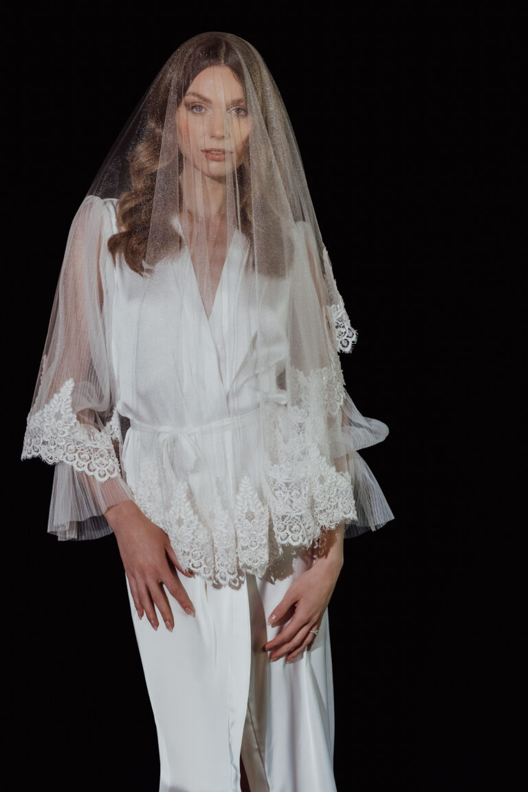 Eine Frau in einem weißen Brautkleid mit Spitzendetails und einem durchsichtigen Schleier vor einem schwarzen Hintergrund - die perfekte Inspiration für Ihre Hochzeitsplanung in der Schweiz mit Hilfe eines erfahrenen Hochzeitsplaners.