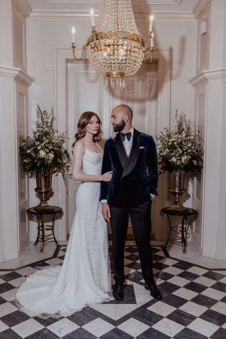 Die Braut im weißen Kleid und der Bräutigam im Samt-Smoking stehen gemeinsam unter einem Kronleuchter in einem eleganten Flur und zeigen eine exquisite Hochzeitsplanung, die vom raffinierten Stil der Schweiz inspiriert ist.