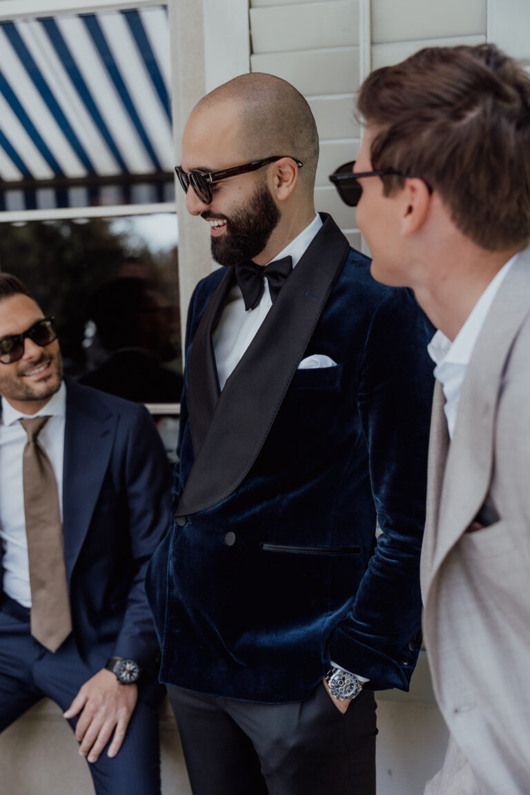 Drei Männer in Anzügen und Sonnenbrillen lächeln und unterhalten sich draußen an einem Fenster mit einer blau-weiß gestreiften Markise über ihre neueste italienische Hochzeitsplanung.