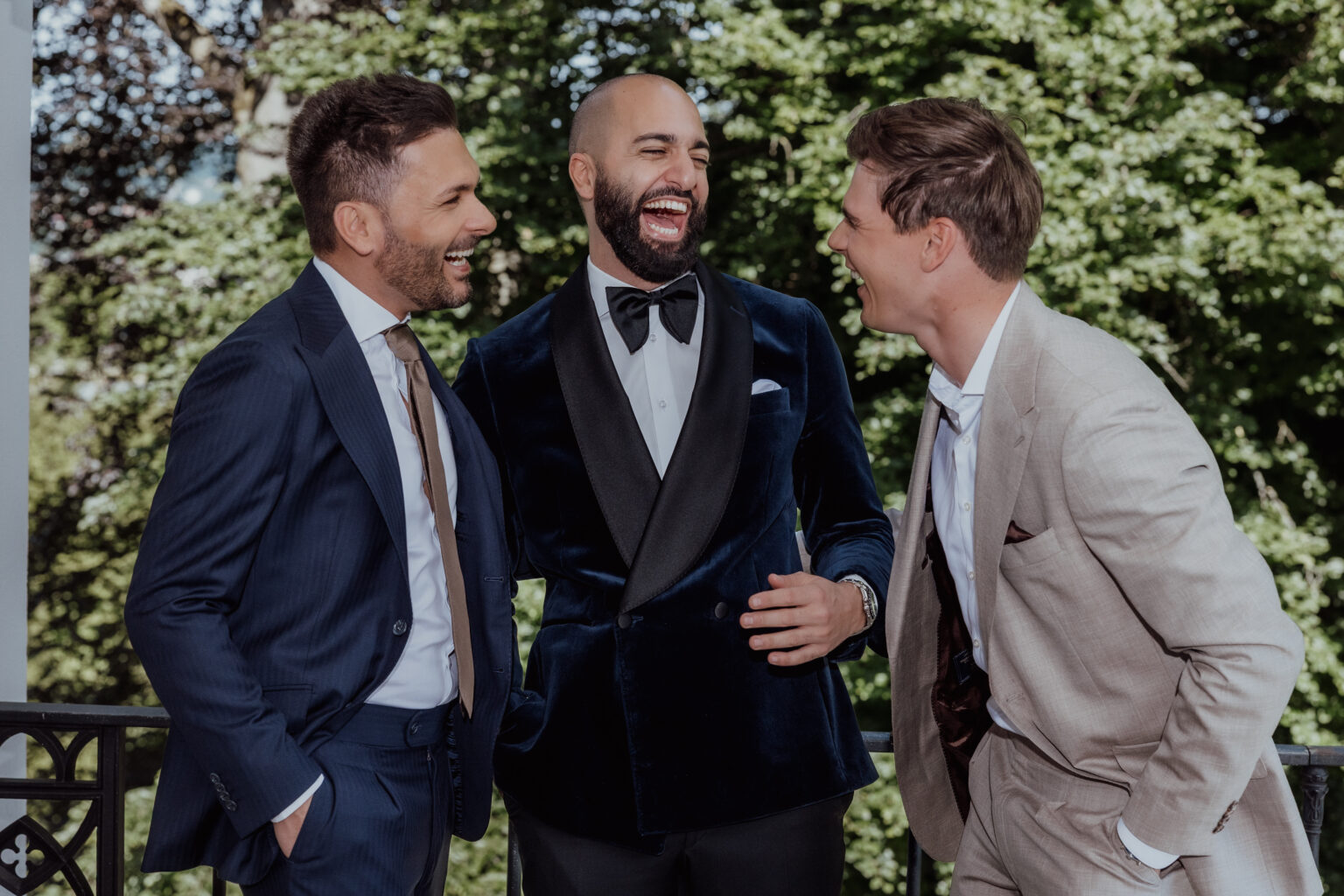 Home 13 Drei Männer in Anzügen, die gemeinsam im Freien vor grünen Bäumen lachen - die perfekte Inspiration für alle, die Hochzeitsplanung betreiben oder einen talentierten Wedding Planner in der Schweiz suchen.