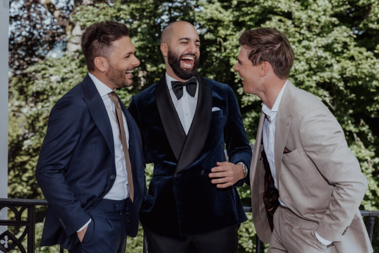 Drei Männer in Anzügen, die gemeinsam im Freien vor grünen Bäumen lachen - die perfekte Inspiration für alle, die Hochzeitsplanung betreiben oder einen talentierten Wedding Planner in der Schweiz suchen.