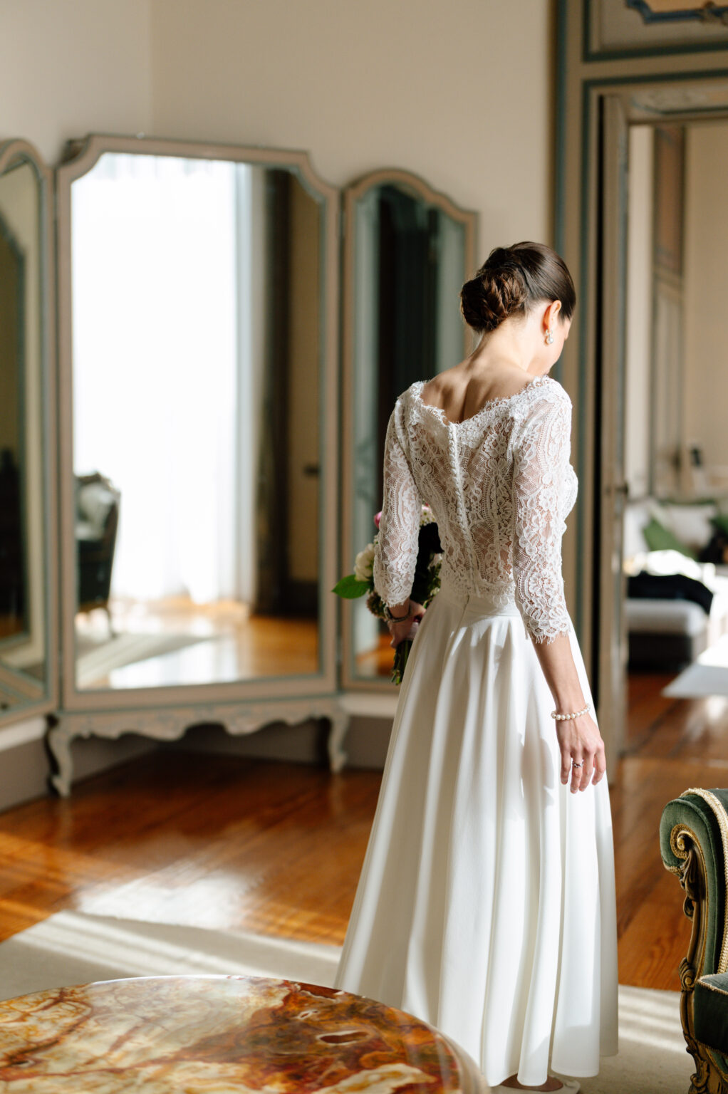 Home 27 Eine Braut im Spitzenhochzeitskleid steht in einem sonnendurchfluteten Raum und hält einen Blumenstrauß in der Hand - die perfekte Inspiration für Ihre Hochzeitsplanung in der Schweiz.