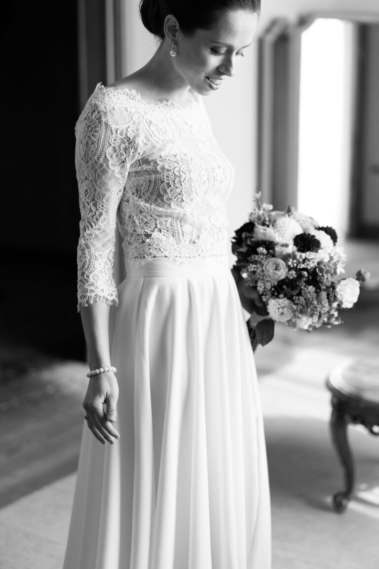 Braut im Spitzenkleid mit Blumenstrauß, lächelnd und nach unten blickend, in einem sanft beleuchteten Raum. Schwarz-Weiß-Foto von einem Hochzeitsplaner, der sich auf elegante Hochzeiten in der Schweiz und Italien spezialisiert hat.
