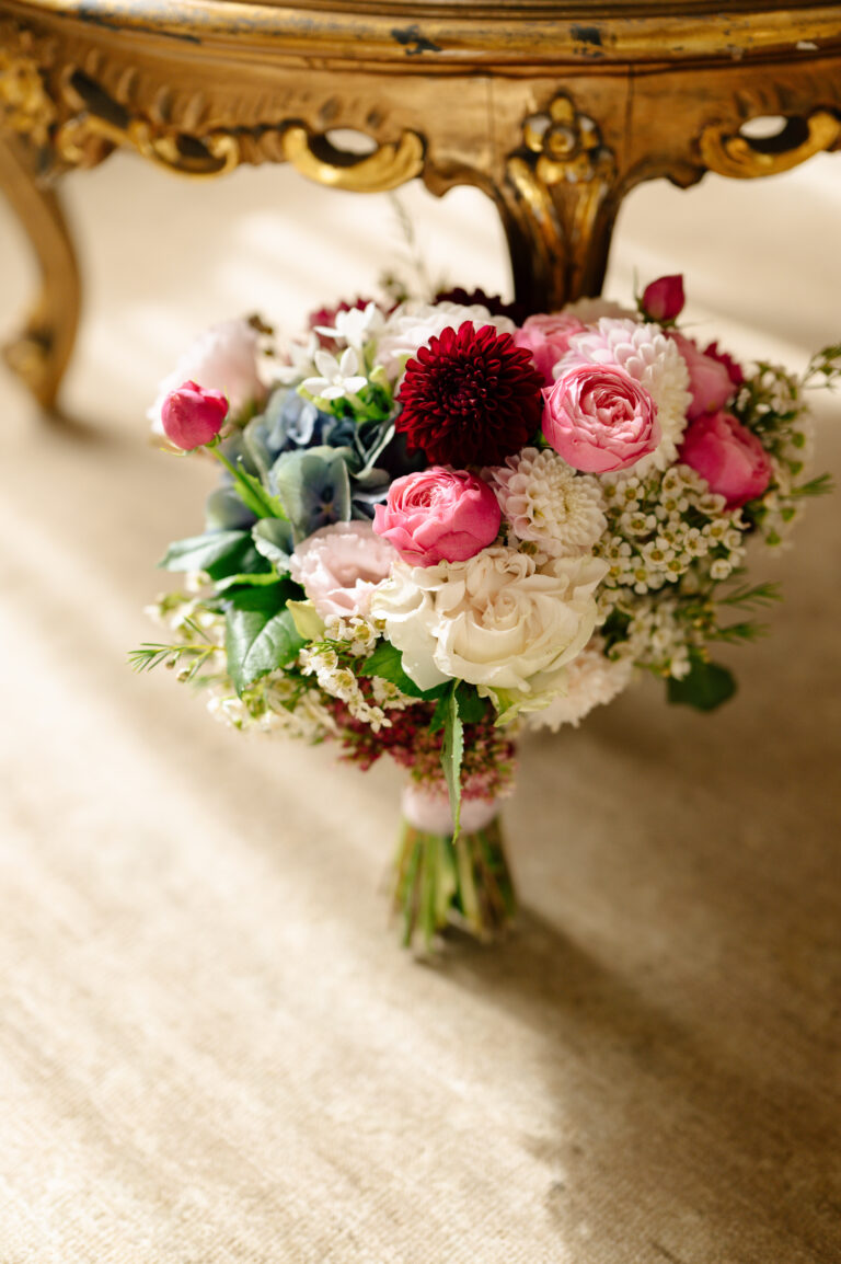 Ein Blumenstrauß aus rosa, roten, weißen und violetten Blumen auf einem beigen Teppich unter einem dekorativen Holztisch - die perfekte Inspiration für jeden Hochzeitsplaner in Italien oder der Schweiz.