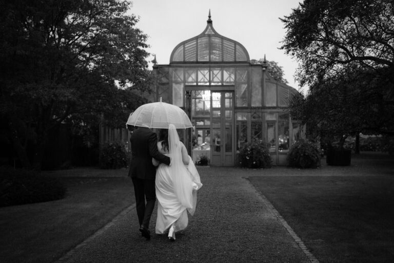 Unter der Anleitung ihres Hochzeitsplaners gehen die Braut und der Bräutigam an einem regnerischen Tag unter einem Regenschirm in Richtung eines gläsernen Wintergartens.