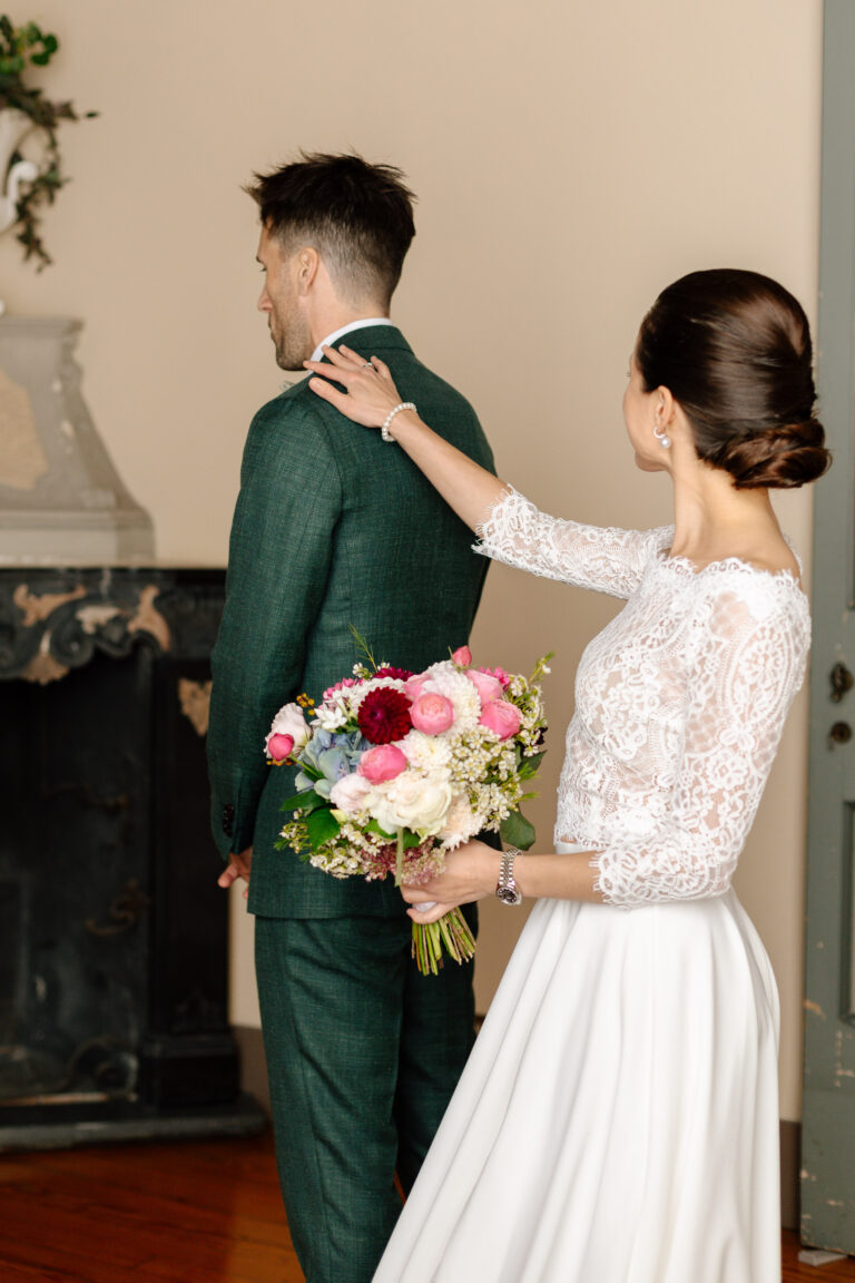 Eine Braut in einem Spitzenkleid berührt die Schulter des Bräutigams, der sich mit einem bunten Blumenstrauß in der Hand abwendet - ein intimer Moment, der von einem professionellen Hochzeitsplaner wunderschön inszeniert wurde.