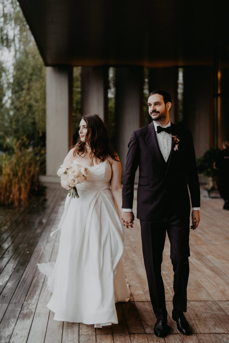 Die Braut im weißen Kleid und der Bräutigam im dunklen Anzug halten sich an den Händen und gehen auf einer Holzterrasse spazieren - die perfekte Inspiration für Ihre italienische Hochzeitsplanung mit Ihrem Wedding Planner.
