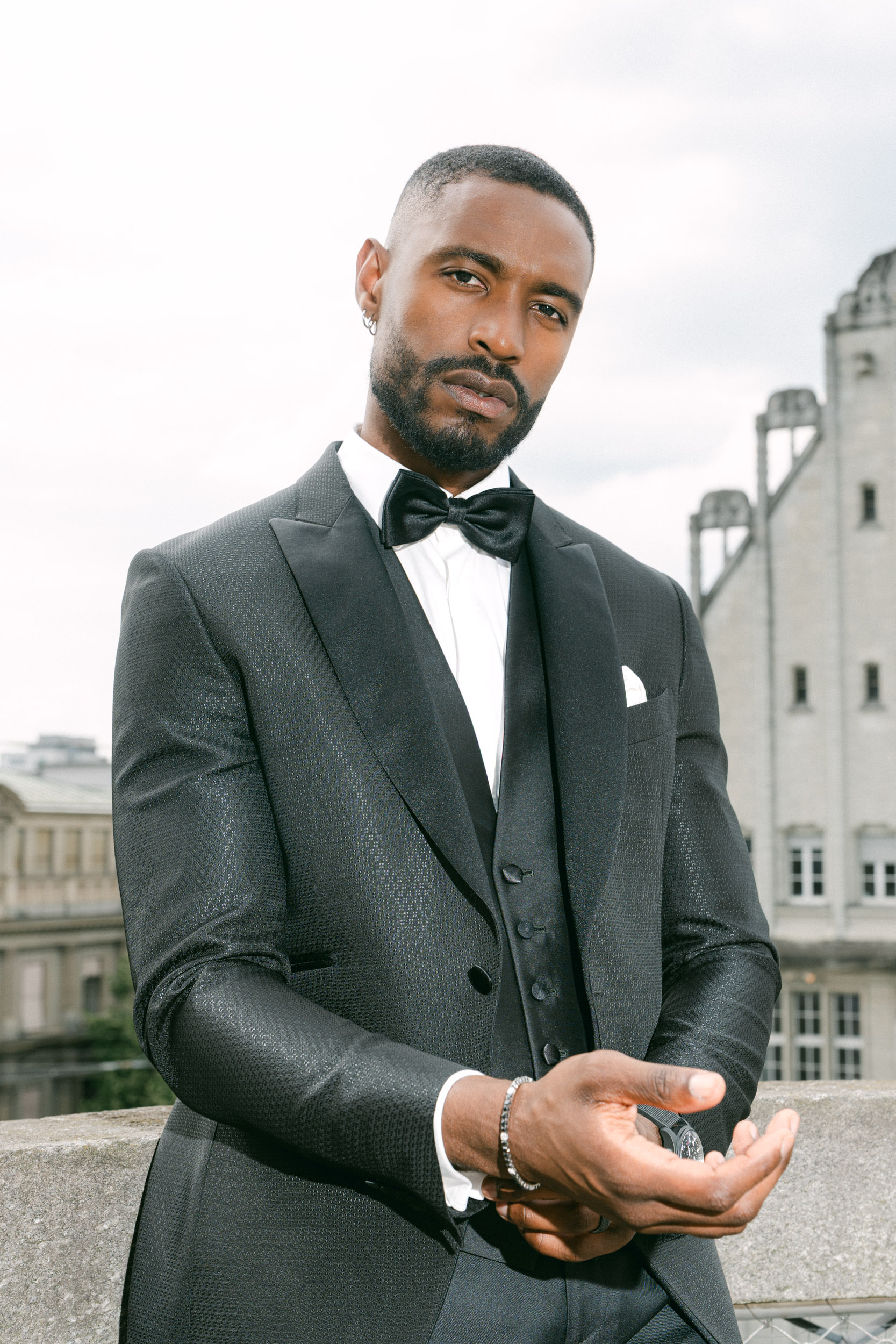 Ein Mann im schwarzen Smoking mit Fliege steht im Freien und posiert selbstbewusst mit Gebäuden im Hintergrund - die perfekte Inspiration für jeden Hochzeitsplaner oder Wedding Planner in der Schweiz.
