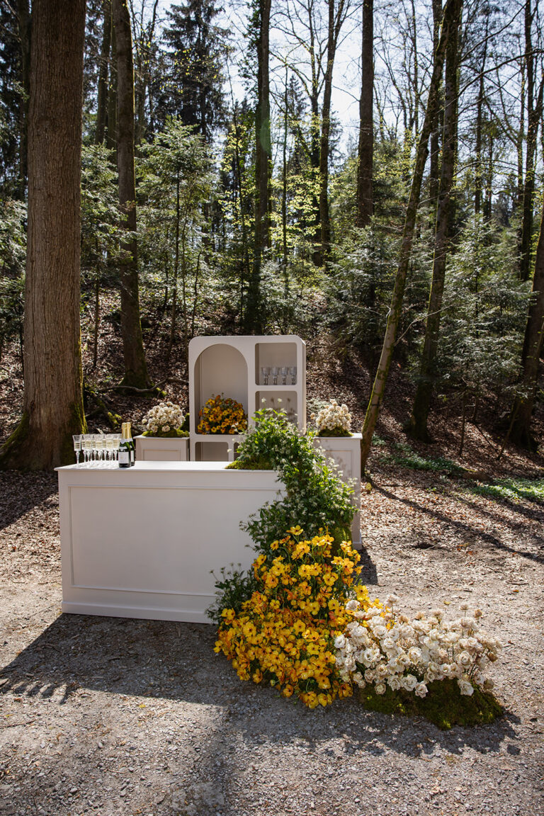 Eine weiße Bar im Freien in einem Wald, dekoriert mit gelben und weißen Blumen, die kaskadenartig zu Boden fallen - die perfekte Inspiration für Ihre Hochzeitsplanung oder wenn Sie mit einem Wedding Planner in Italien zusammenarbeiten.