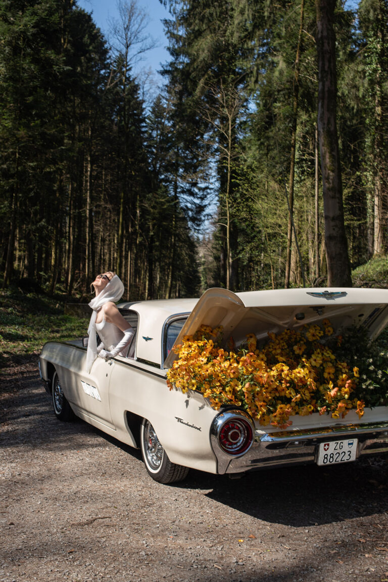 Eine Frau in einem weißen Kleid lehnt sich aus einem mit gelben Blumen gefüllten Oldtimer in einem Wald - eine bezaubernde Szene für jeden Hochzeitsplaner, der Inspiration sucht.
