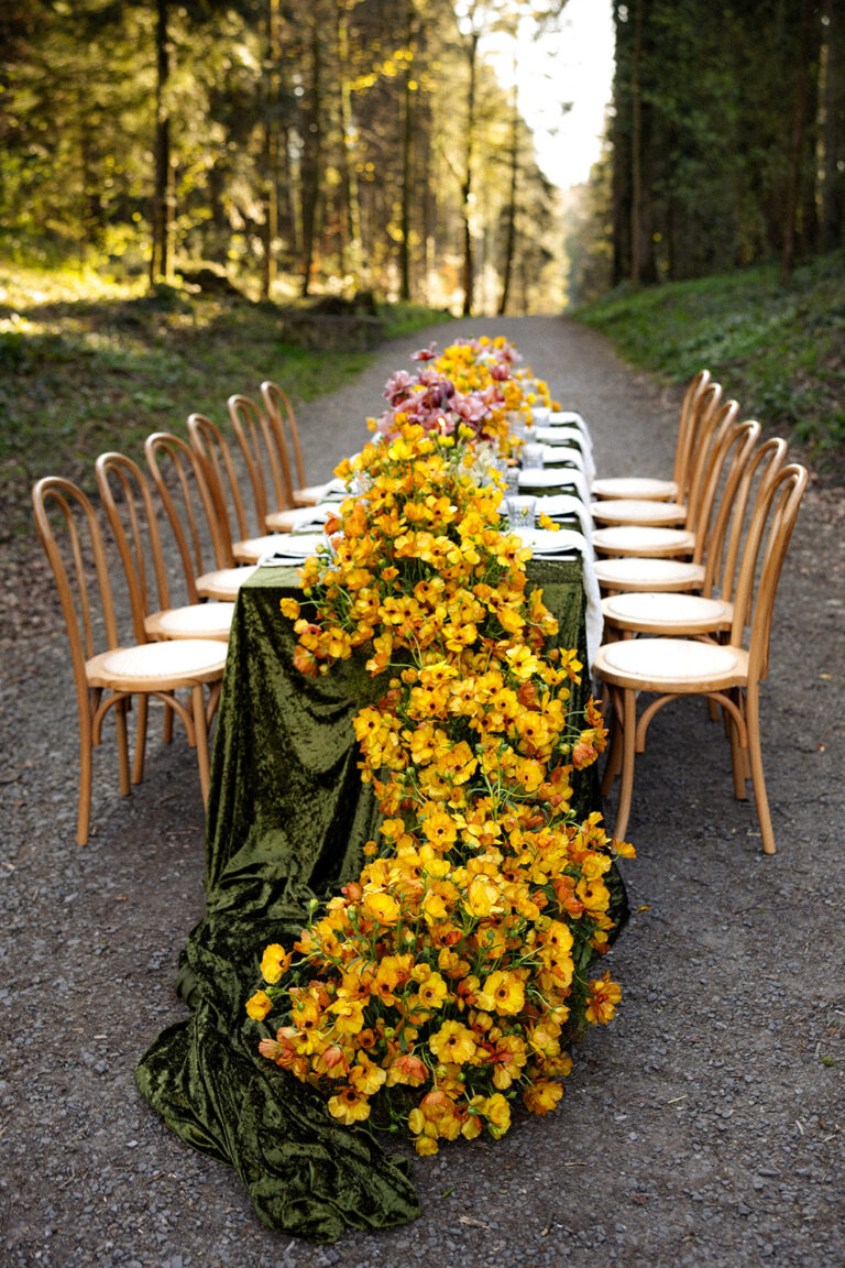 Ein langer Esstisch im Freien an einem Waldweg, dekoriert mit gelben und orangefarbenen Blumen - die perfekte Inspiration für alle Hochzeitsplaner, die eine zauberhafte, von der Natur inspirierte Kulisse suchen.