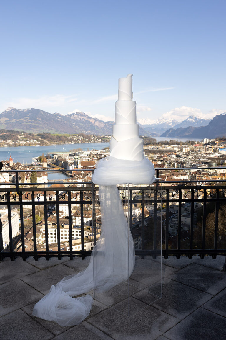 Eine hohe weiße Hochzeitstorte mit einem langen Tüllschleier steht auf einer Terrasse mit Blick auf eine Stadt und schneebedeckte Berge, perfekt für eine Schweizer oder italienische Feier, die von einem erfahrenen Wedding Planner geplant wurde.