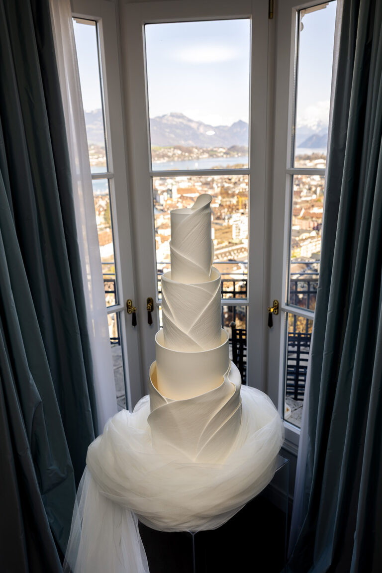 Hohe, elegante weiße Hochzeitstorte mit spiralförmigen Schichten an einem Fenster mit Blick auf die Stadt und die Berge - perfekt für eine italienisch inspirierte Feier, entworfen von Ihrem Hochzeitsplaner.
