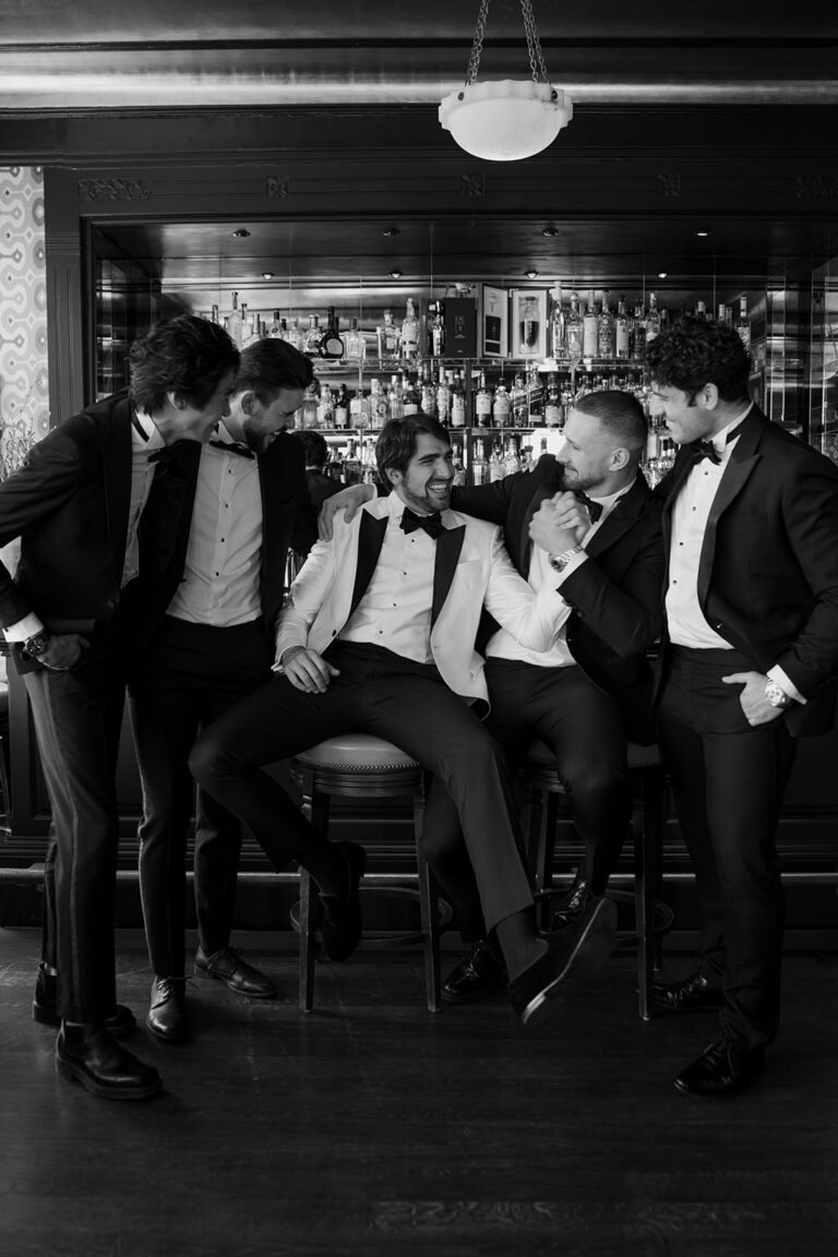 Fünf Männer im Smoking, lächelnd und im Gespräch vor einer Bar - einer sitzend, die anderen stehend - genießen einen stilvollen Moment, vielleicht besprechen sie die Hochzeitsplanung mit ihrem Wedding Planner vor einer Traumveranstaltung in Italien.