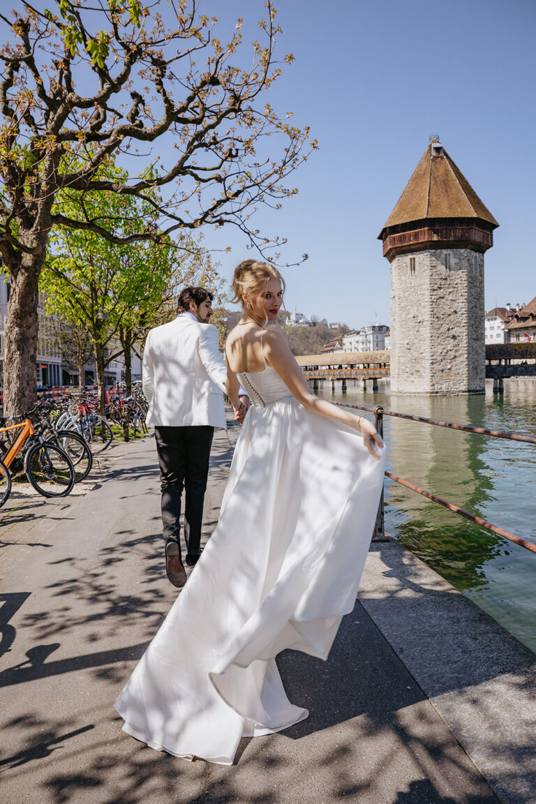 Die Braut im weißen Kleid lächelt zurück, während sie mit dem Bräutigam am Flussufer in der Nähe eines historischen Steinturms spazieren geht - ein unvergesslicher Moment, der von ihrem Hochzeitsplaner festgehalten wurde.