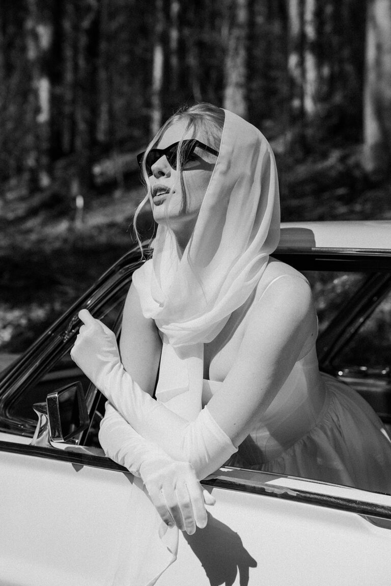 Eine Frau mit Sonnenbrille und Schal lehnt sich mit Handschuhen aus einem Autofenster. Ein Schwarz-Weiß-Foto, das die stilvolle Hochzeitsplanung in der Schweiz perfekt inspiriert.