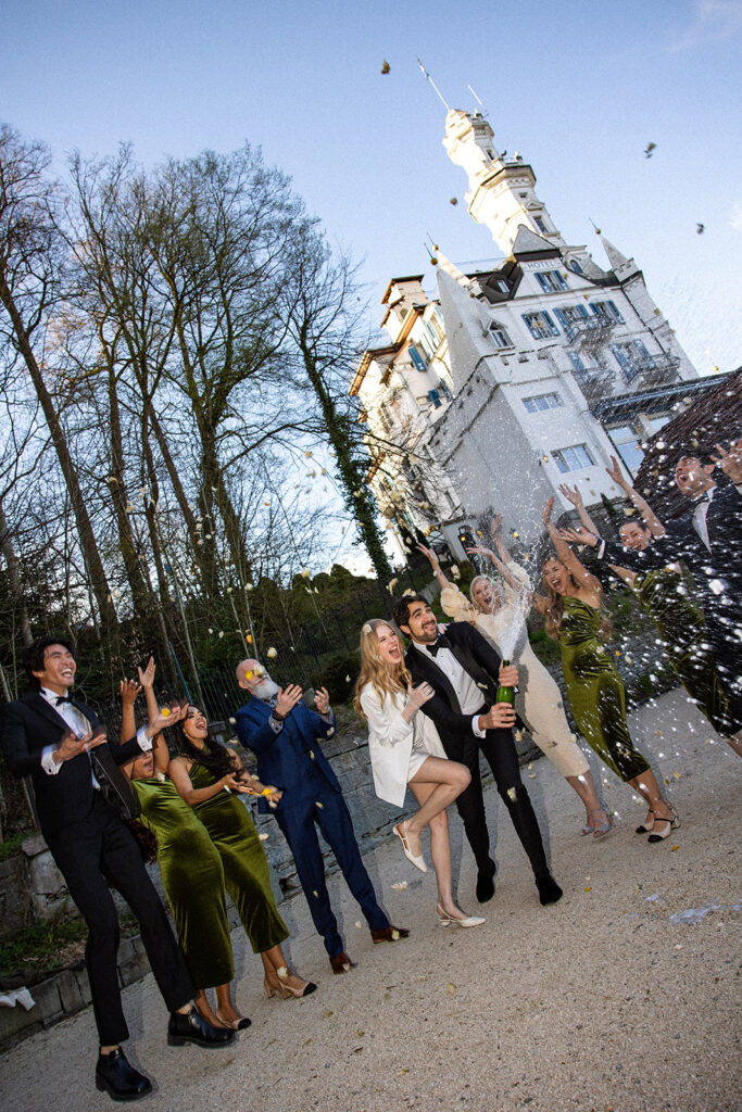 Eine Gruppe förmlich gekleideter Menschen feiert in der Schweiz im Freien, lässt Sekt knallen und jubelt in der Nähe eines weißen Gebäudes - die perfekte Inspiration für die Hochzeitsplanung durch einen erfahrenen Hochzeitsplaner.