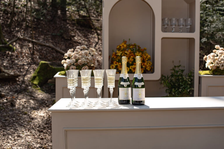 Vier Gläser und zwei Flaschen Champagner auf einer weißen Bar im Freien, mit Blumen im Hintergrund - die perfekte Inspiration für die Hochzeitsplanung oder jeden Wedding Planner in der Schweiz.