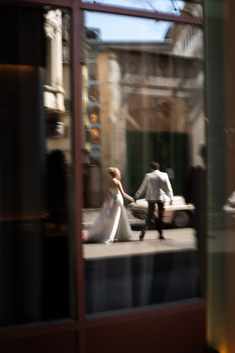 Eine Braut und ein Bräutigam halten sich an den Händen, während sie durch eine spiegelnde Glasscheibe auf einer Straße in der Schweiz spazieren gehen - ein Bild, von dem jeder Hochzeitsplaner träumt.