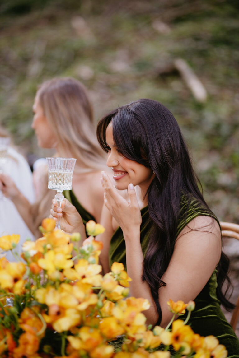 Eine Frau in einem grünen Kleid, lächelnd und mit einem Glas in der Hand, sitzt zwischen gelben Blumen bei einem Treffen im Freien - die perfekte Inspiration für jede Hochzeitsplanung in der Schweiz.