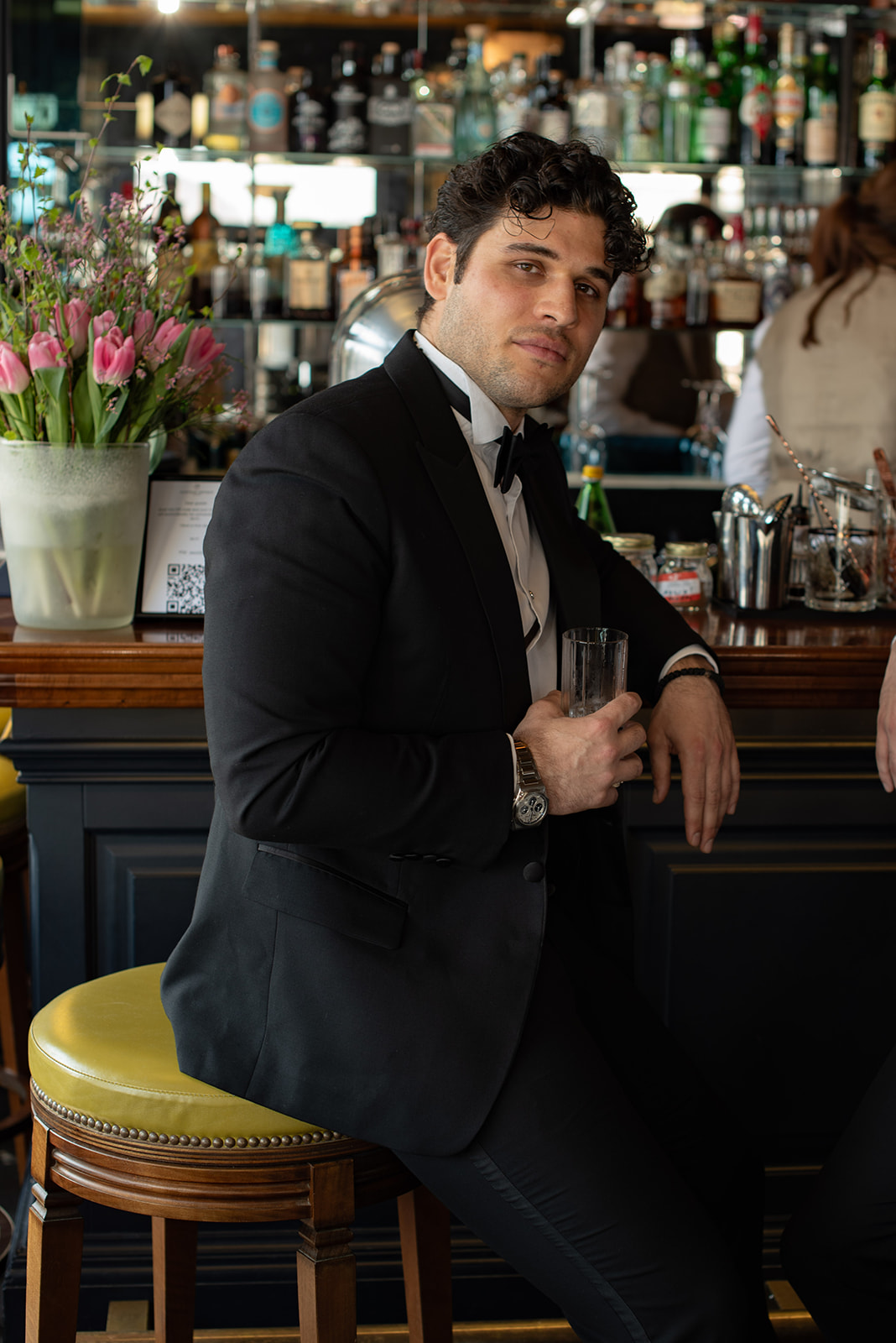 Ein Mann im schwarzen Smoking sitzt an einer Bar und hält einen Drink in der Hand. Im Hintergrund sind Blumen und Flaschen zu sehen - die perfekte Inspiration für jeden Wedding Planner, der an der Hochzeitsplanung in der Schweiz arbeitet.