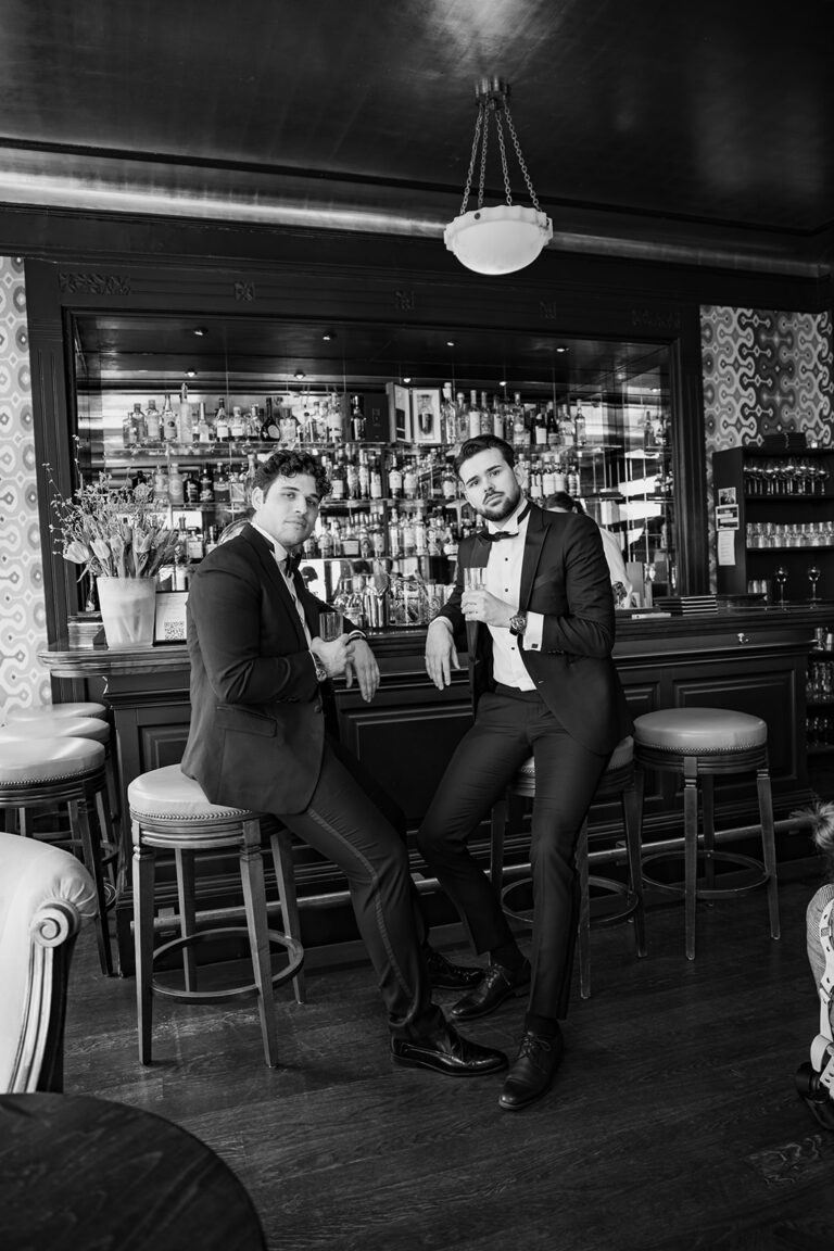 Zwei Männer in Anzügen sitzen an einer Bar, halten Drinks in der Hand und blicken in die Kamera. Eine stilvolle, elegante Szenerie - perfekt für einen Hochzeitsplaner, der nach Inspiration für anspruchsvolle Feiern in der Schweiz oder Italien sucht.